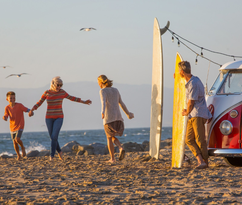 Family running together on a beach with surfboards stuck in sand, gulls overhead and a two-tone red and white T1 Volkswagen bus off to the side.